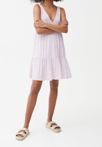 Helllila, ärmelloses Kleid mit V-Ausschnitt, floralem Muster und einem gestuften Rock. Getragen mit beigen Sandalen mit breiten Riemen.
