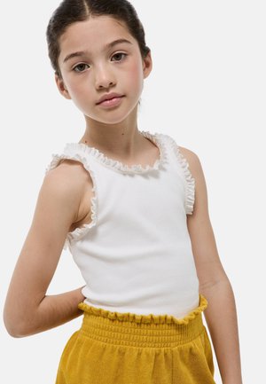 Jeune fille aux cheveux foncés tirés en arrière, portant un haut blanc sans manches à volants et un pantalon jaune moutarde texturé, debout devant un fond clair.