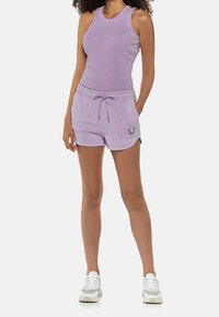 Sada fialkového topu na ramínka a šortek vyrobená z měkké tkaniny, s pasem na šňůrku a akcentem loga na šortkách. Tenisky dotvářejí celý outfit.