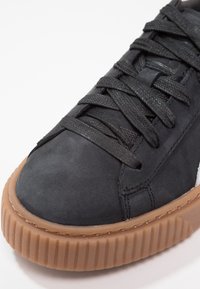 Gros plan sur une basket noire avec une semelle épaisse marron à rainures et des lacets noirs, mettant en valeur les matériaux texturés et les détails de couture.