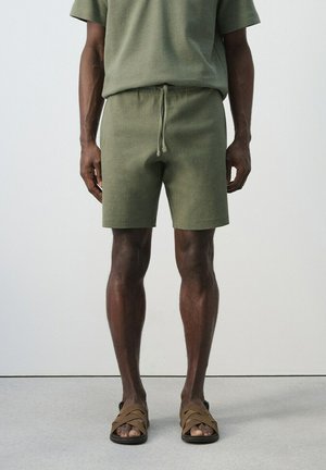 Uomo che indossa pantaloncini in maglia verde oliva e maglietta a maniche corte abbinate con sandali con cinturino marrone, in piedi su un pavimento grigio chiaro contro una parete liscia.