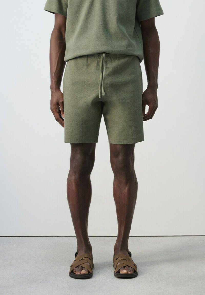 Homme portant un short en maille vert olive et un haut à manches courtes assorti, avec des sandales à lanières marron, debout sur un sol gris clair devant un mur uni.