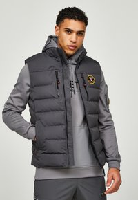 Grijze gewatteerde vest met hoge kraag, voorzien van een ritssluiting aan de voorkant, twee ritszakken en een logo label aan de linkerkant.