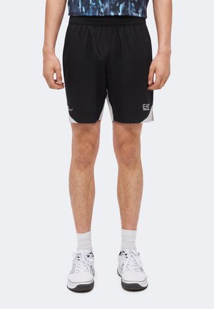 Homme portant un short de sport noir et gris, des chaussettes blanches et des chaussures de sport blanches, debout face à un fond uni.