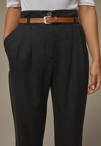 Crne hlače opuštenog kroja, s visokim strukom, bočnim džepovima i smešnim kožnim pojasom s metalnom kopčom. Glatka tekstura.