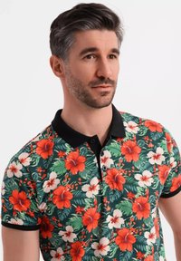 Kortärmad polo t-shirt med ett livligt blommönster av röda hibiskusblommor och vita blommor på en grön bakgrund, svart krage och knappar.