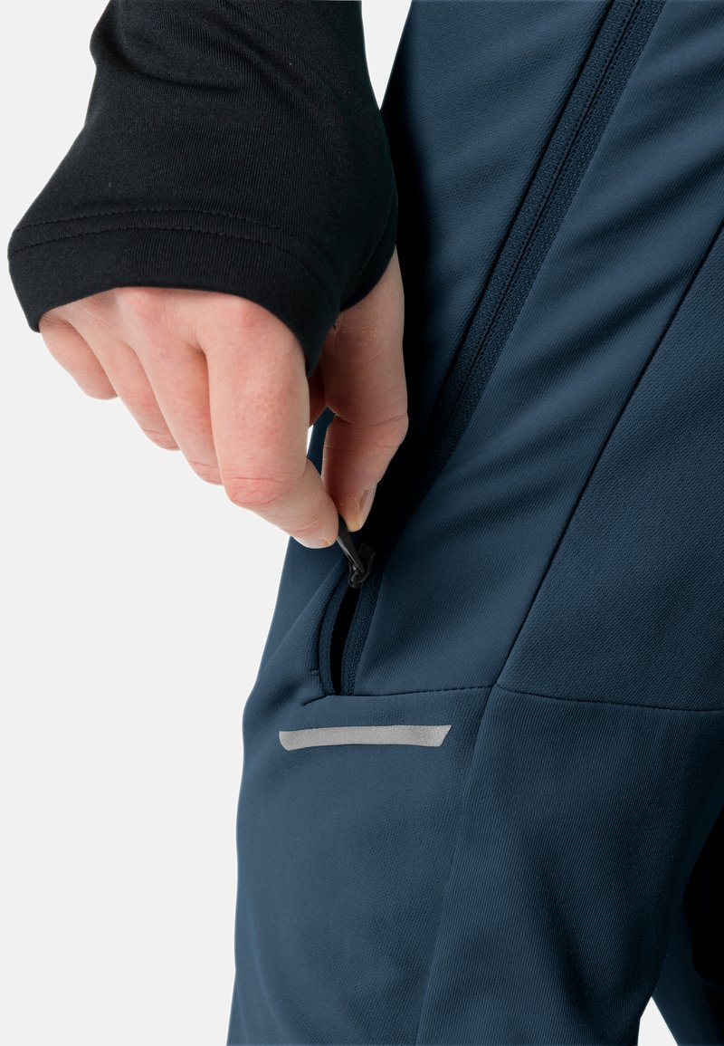 Hand mit schwarzem Ärmel, die den Reißverschluss der Seitentasche eines dunkelblauen Stoffkleidungsstücks mit reflektierendem Streifen zieht.