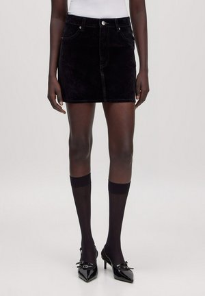 Vrouw die een korte zwarte corduroy rok draagt, zwarte kniehoge doorschijnende sokken en zwarte puntige hakken met gespen.