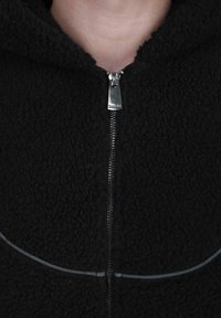 Giacca in pile nera con zip fino al collo, caratterizzata da un evidente tirazip in argento e dettagli con cuciture curve vicino al petto.