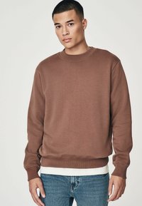 Sudadera marrón con cuello redondo, puños acanalados y dobladillo. Lleva puesta una camiseta blanca debajo y se combina con jeans azules. Tejido suave y texturizado.