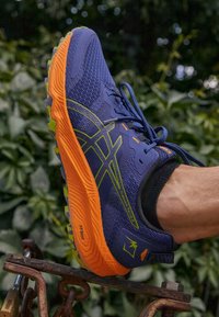 Männlicher Laufschuh aus blauem Mesh mit grünen Akzenten, orangefarbiger Zwischensohle, strukturiertem Sohlendesign und schwarzem Schaft. Verfügt über Logo- und Markendetails.