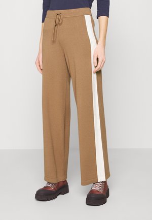 Persoon draagt bruine wijdere broek met witte zijstrepen en bruine chunky laarzen, staand tegen een effen lichte achtergrond.