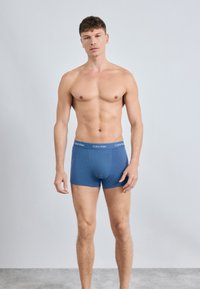 Mėlyni medvilniniai boxer tipo apatiniai su logotipo juosta, pasižymintys prigludusia forma, lygiu paviršiumi ir vidutinio ilgio iki šlaunų. Modelis stovi prieš paprastą foną.