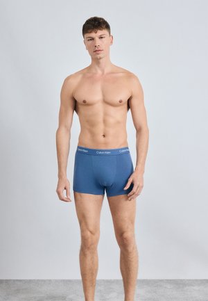 Blue cotton boxer briefs with a logo band, featuring a fitted design, smooth texture, and mid-thigh length. Model stands against a plain background.