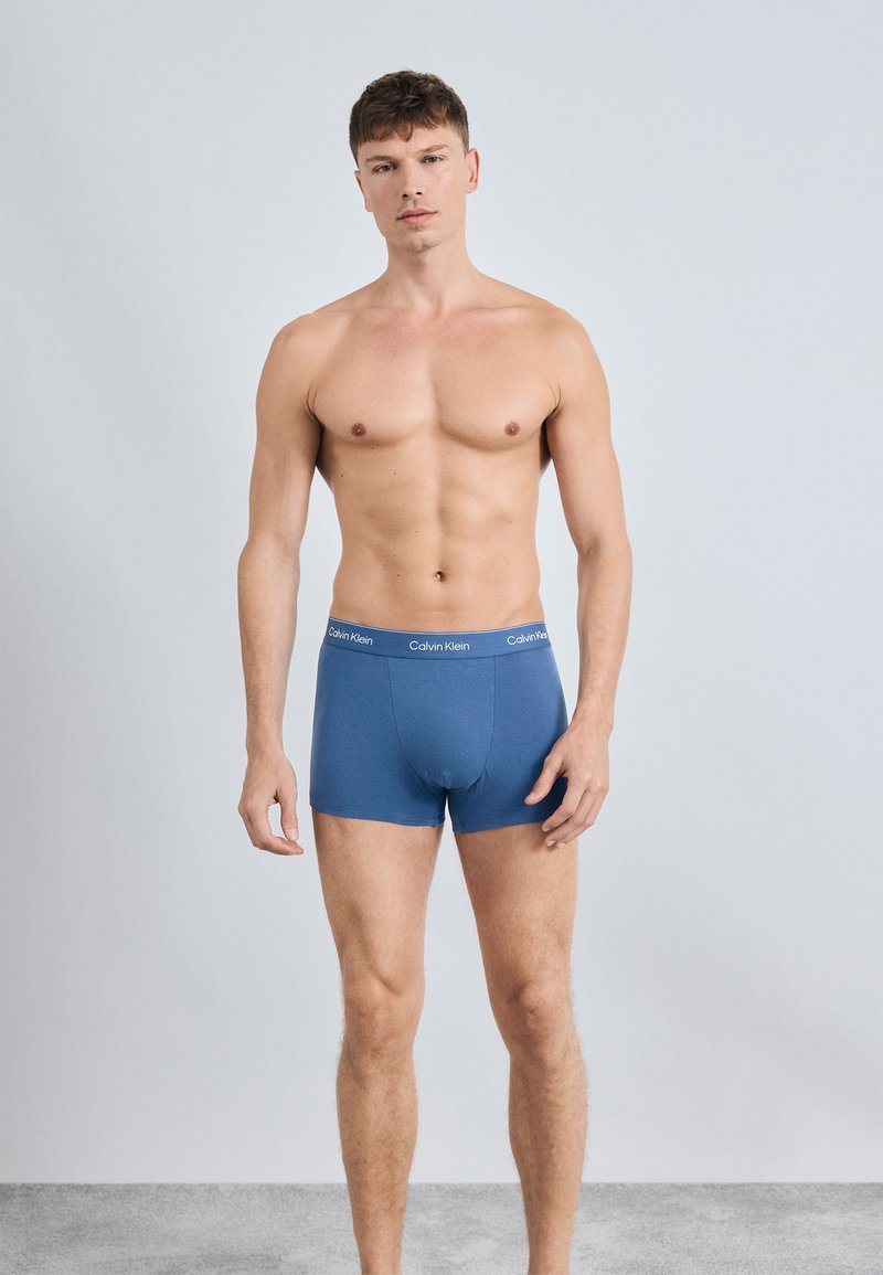 Mėlyni medvilniniai boxer tipo apatiniai su logotipo juosta, pasižymintys prigludusia forma, lygiu paviršiumi ir vidutinio ilgio iki šlaunų. Modelis stovi prieš paprastą foną.