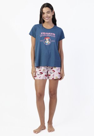 Joven sonriente que lleva una camiseta azul con el gráfico "Jessie" y pantalones cortos rosa claro con patrones coloridos inspirados en Toy Story, descalza.