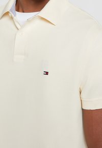 Kräme polo skjorta med korta ärmar, med en klassisk krage och en liten broderad logotyp i blått, rött och vitt på bröstet.