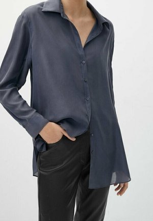 Femme portant un chemisier ample bleu foncé à boutons avec manches longues et un pantalon noir, une main dans la poche.
