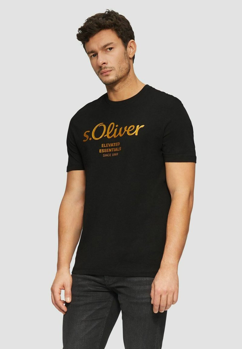 Black cotton t-shirt featuring gold printed text "s.Oliver" and "Elevated Essentials Since 1969." Short sleeves and crew neckline.