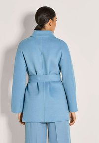 Vrouw met donker haar in een lage paardenstaart, gekleed in een lichtblauwe mantel met riem en bijpassende wijde broek, met haar rug naar de camera toe tegen een effen achtergrond.