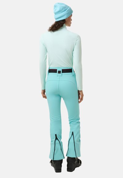 Mintgrünes Skitop und passende Hose mit Reißverschlüssen an den Knöcheln, ausgestattet mit einem schwarzen Gürtel. Das Modell trägt eine hellblaue Mütze.