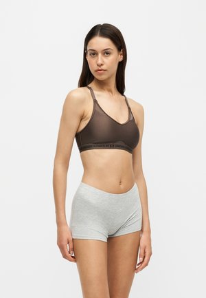Junge Frau steht und schaut nach vorne, trägt einen braunen Sport-BH und hellgraue Shorts vor einem einfarbigen weißen Hintergrund.