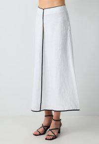Jupe maxi en lin blanc avec une bordure noire le long des coutures et une silhouette droite. Présente une fermeture éclair sur le côté et un design minimaliste.