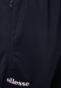 Joggers de color navy hechos de un tejido texturizado que cuentan con un bolsillo con cremallera y un logo bordado en blanco y negro de "ellesse" en la pierna.
