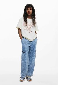 Krém színű póló piros felirattal, világoskék cargo nadrággal, melyen finom virágminták találhatók, és ezüst sportcipővel párosítva.