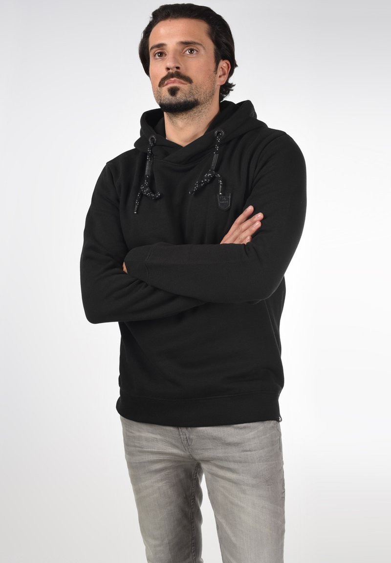 Sweatshirt à capuche noir avec des cordons texturés, détail du logo sur la poitrine et poignets côtelés. Porté avec un jean gris clair.