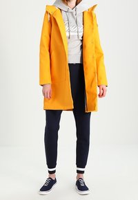 Geel waterdicht regenjack met een capuchon, een grijze hoodie eronder, donkerblauwe broek en marineblauwe sneakers. Bevat drukknopen en zakken.