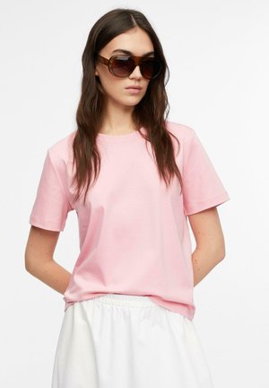 Kobieta z długimi brązowymi włosami, nosząca za duże brązowe okulary przeciwsłoneczne, jasnoróżowy t-shirt i białe spodnie z elastycznym pasem.