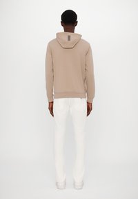 Sudadera con capucha beige y cordón, con un detalle de logotipo en la parte trasera, combinada con jeans ajustados blancos y zapatillas ligeras.