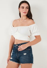 Hvid off-the-shoulder top med sammensynkede ærmer, parret med mørkeblå denimshorts med frynset kant og let slitage.