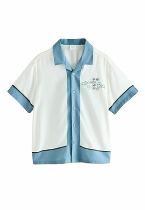 Jongen in korte broek en sandalen loopt langs een stenen richel naast een witte, door de zon verlichte muur die schaduwen werpt.