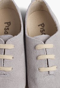 Chaussures en suède gris clair avec une surface texturée, dotées de trois lacets de couleur crème et de détails de couture subtils. L'intérieur présente le nom de la marque.