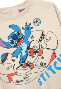 Felpa beige con un personaggio dei cartoni animati blu su una tavola da skateboard, con stampe vivaci di pianeti e disegni in vari colori.