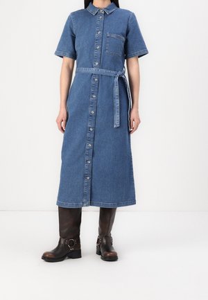 Femme portant une robe en denim bleu mi-longue à manches courtes avec des boutons et une ceinture à la taille, associée à des bottes hautes marron foncé jusqu'aux genoux.