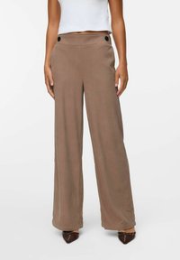 Un pantalon large en tissu ribbed marron doux. Dispose d'une taille haute, d'une fermeture à l'avant et de deux grands boutons noirs au niveau de la taille.