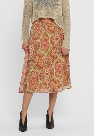 Eine Person steht mit einem beigen Strickoberteil und einem gemusterten Midikleid mit orangefarbenen, pinken und grünen Motiven, kombiniert mit schwarzen Ankle Boots.