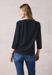 Musta teksturoitu paita, jossa on kolme neljännesosaa hihat, rento istuvuus ja pudotettu helma; paritettu tiukasti istuvien sinisten farkkujen kanssa.