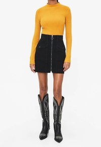 Senf-farbenes geripptes Langarmoberteil, schwarzer Denim-Minirock mit Vorderreißverschluss und Taschen, kombiniert mit kniehohen schwarzen Cowboy-Stiefeln mit silbernen Details.