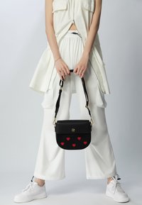 Černá kožená kabelka s aplikacemi červeného srdce, zlatými doplňky a nastavitelným popruhem, v kombinaci s bílým oversized oblečením a teniskami.