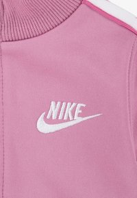 Rosa strikket stoff med høy krage, hvit brodert Nike-logo og kontrasterende skulderdetaljer i hvitt og rosa.