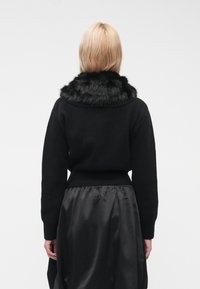 Pulover negru cu guler moale din blană sintetică, manșete striate și talie ajustată, suprapus peste o fustă neagră lucioasă. Vedere din spate.