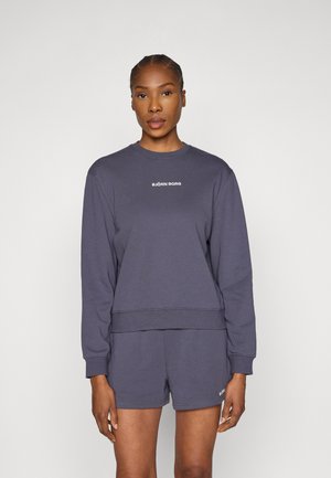 Siva majica z dolgimi rokavi, rebrastimi manšetami in ravnim robom. Na prsih je bel logotip. Kombinirana z usklajenimi kratkimi hlačami.