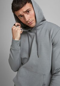 Sudadera gris con cordón y bolsillo frontal, con una textura suave y puños acanalados. El modelo presenta un perfil medio con una expresión seria.