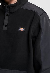 Jersey de forro polar negro con hombros de tela oscura, cierre con botones a presión y un pequeño parche con el logo de Dickies en el pecho. Textura suave.
