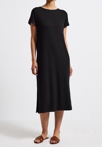 Kobieta w czarnej midi sukience z prążkowanego materiału, z krótkimi rękawami i brązowych płaskich sandałach, stojąca na tle jednolitej białej ściany.