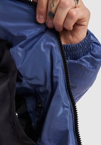 Blå innerfoder i en jacka med en slät textur, elastiska ärmslut och ett svart ytterlager med dragkedja. Hand som greppar öppningen.
